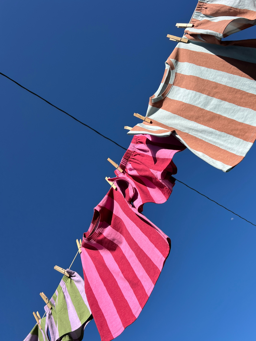Three UMBRELLA STRIPE SHORT SETS, from our unisex matching collection, hang on a clothesline with clothespins beneath a clear blue sky.