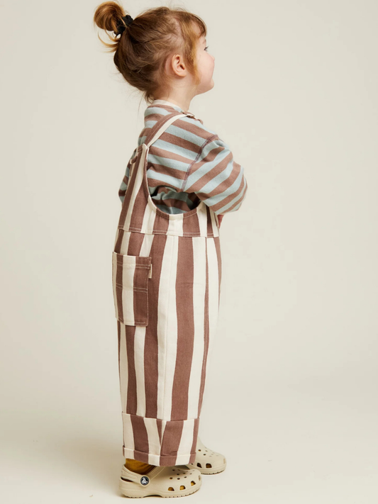 Second image of The BLYTHE CHOC STRIPE DUNGAREE features wide vertical brown and white stripes, adjustable shoulder straps with metal fasteners, a front pocket, and cuffed hems for children.