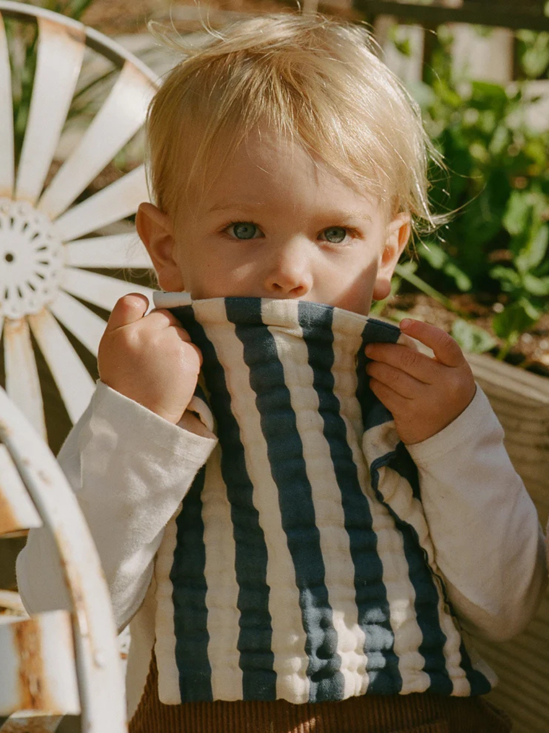 A young child with blonde hair holds an ORGANIC BABY WASH CLOTH up to their mouth while sitting outside on a metal chair.