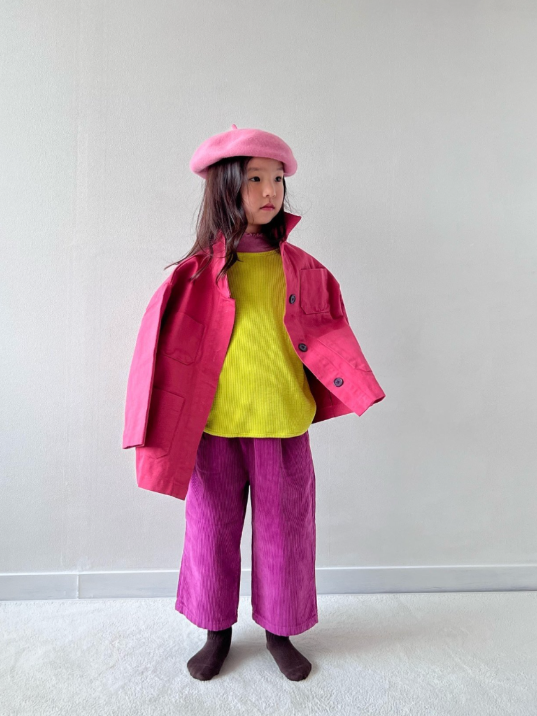 Magenta | A child wearing wide-leg corduroy pants in magenta pink, paired with a bright yellow shirt, pink jacket and light pink beret. She is standing on grey carpet against a white wall..