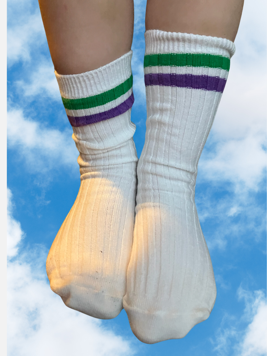 A pair of white ribbed kids socks with purple and green stripes at the top shown word on a kids'd feet, cut out on a background of clouds.