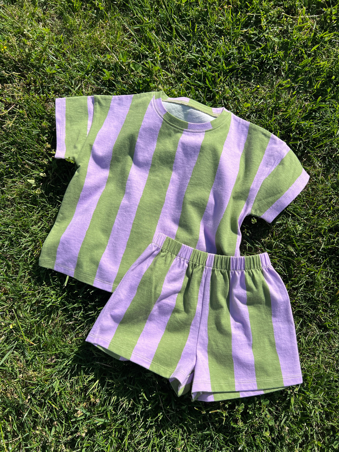 The UMBRELLA STRIPE SHORT SET, a unisex short-sleeve shirt and shorts with bold green and lavender stripes, is displayed on grass—ideal for playful days in stylish striped sets.