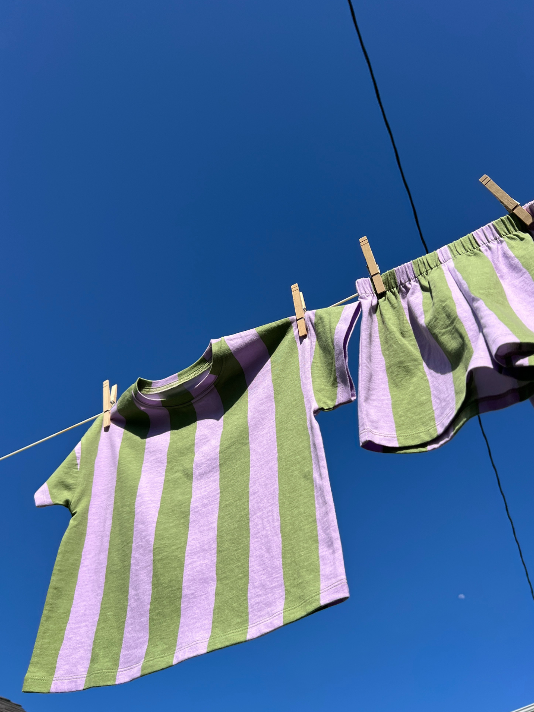 The UMBRELLA STRIPE SHORT SET—a green and lavender striped organic cotton t-shirt and matching shorts—hangs on a clothesline with clothespins under a clear blue sky.