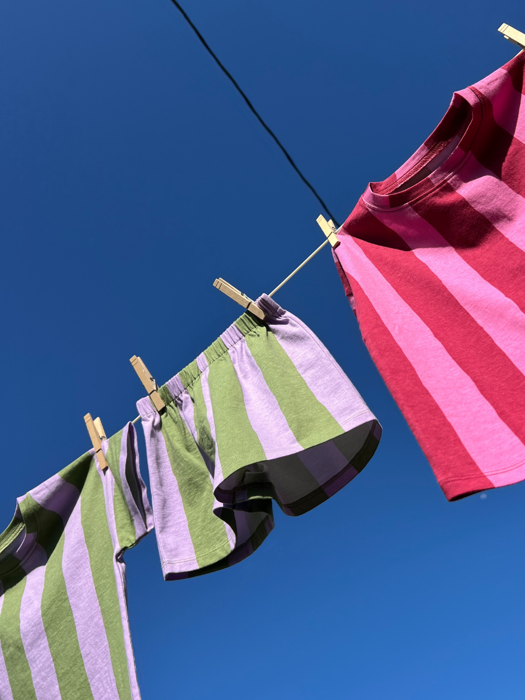 The UMBRELLA STRIPE SHORT SET, made from organic cotton in purple, green, and red stripes, hangs with wooden clothespins on a clothesline beneath a clear blue sky.