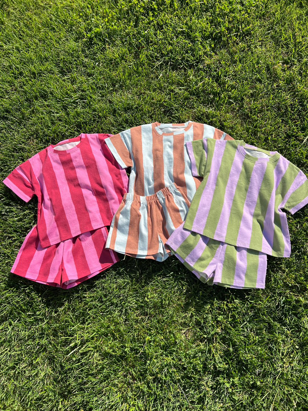 Three unisex UMBRELLA STRIPE SHORT SETS in red-pink, orange-blue, and green-purple striped colorways are displayed on green grass. Each set is made from soft, breathable organic cotton for all-day comfort.