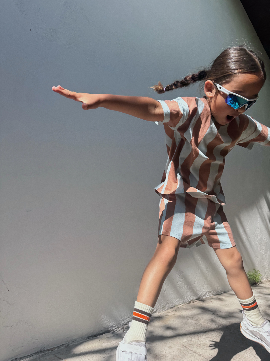 A child in the UMBRELLA STRIPE SHORT SET, sunglasses, and sneakers jumps with arms outstretched against a plain wall in sunlight.