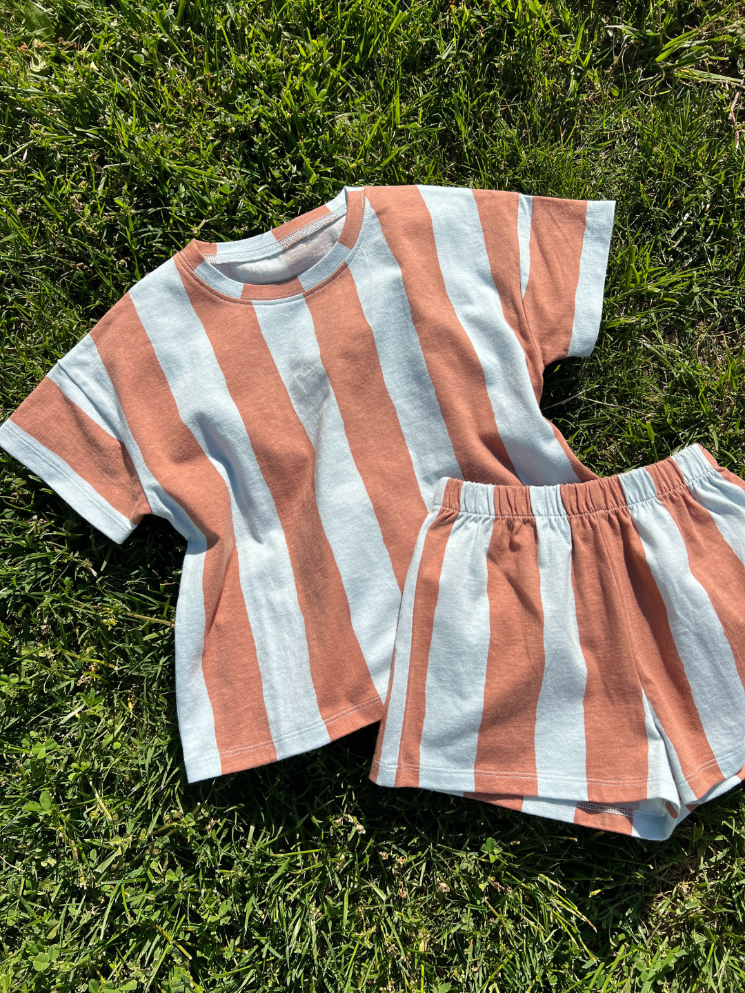 The UMBRELLA STRIPE SHORT SET, made from organic cotton, is a unisex set with a short-sleeve shirt and shorts in bold white and rust-orange stripes, pictured on green grass.