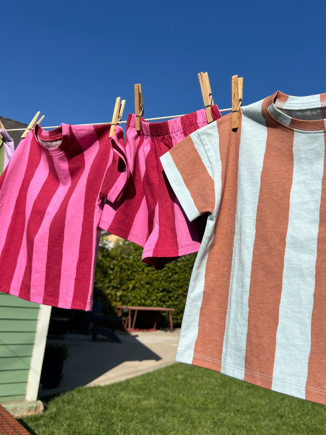 Three UMBRELLA STRIPE SHORT SETS—made from organic cotton, in two pink and one brown and blue—hang on a clothesline outside under a clear blue sky.
