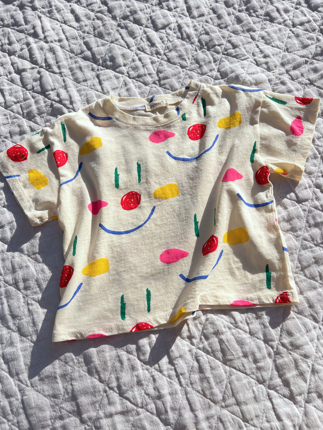 A kids' Facepaint Tee in Cream laid flat on a quilt in the sun