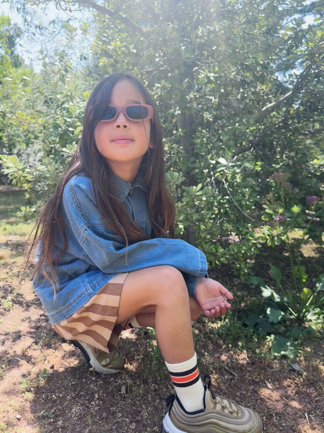 Terracotta | A girl wearing the SANTA CRUZ SUNGLASSES in terracotta, worn with a blue denim jacket, brown striped shorts, and sneakers, kneeling outdoors in a green park.