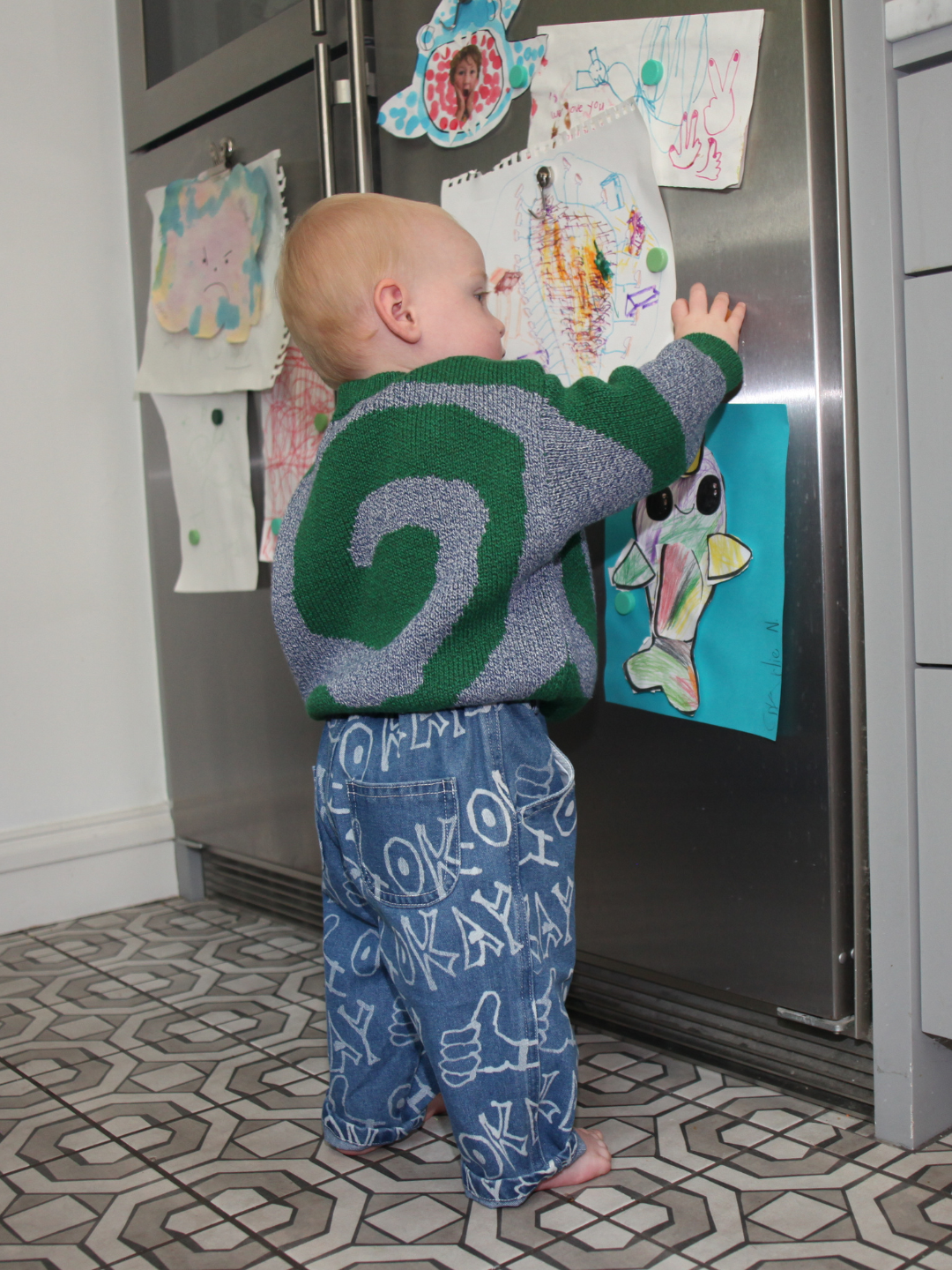 A toddler in OKAY JEANS and a patterned sweater stands on a tiled floor, reaching to touch children's drawings displayed on a refrigerator.