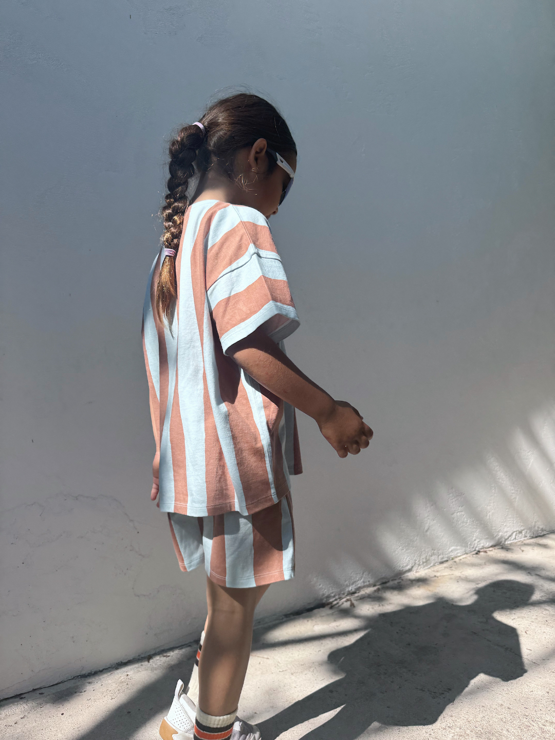 A young girl with braided hair and sunglasses wears the UMBRELLA STRIPE SHORT SET, made from organic cotton, as she stands in sunlight by a white wall.