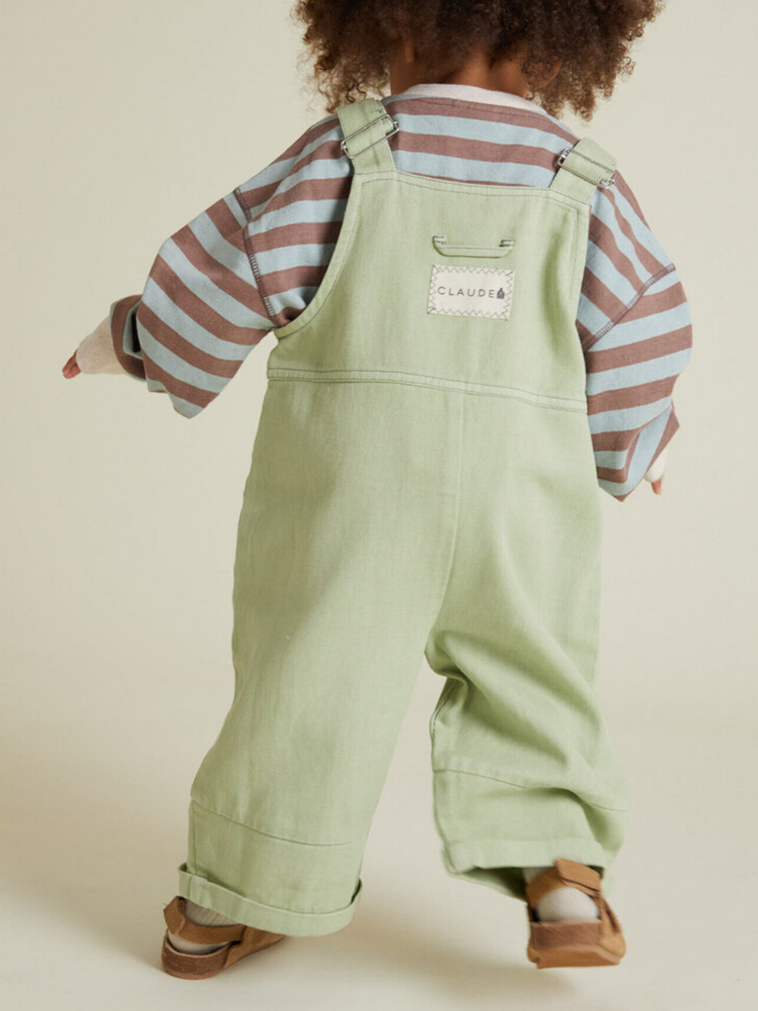 A young child wears the BLYTHE MINT DUNGAREE, paired with a striped long-sleeve shirt and brown shoes, standing with their back to the camera.