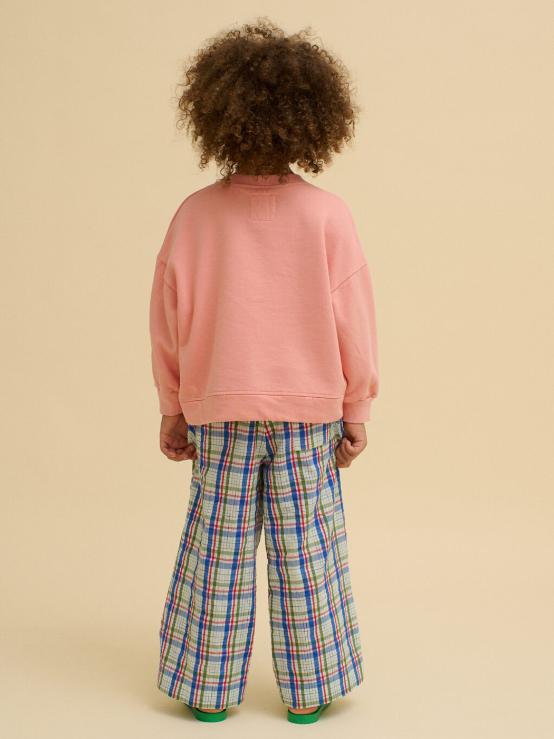 Child with back to camera wears a BOXY POTATO SWEATSHIRT in pink, made from organic cotton, and wide-leg plaid pants, standing against a beige background.