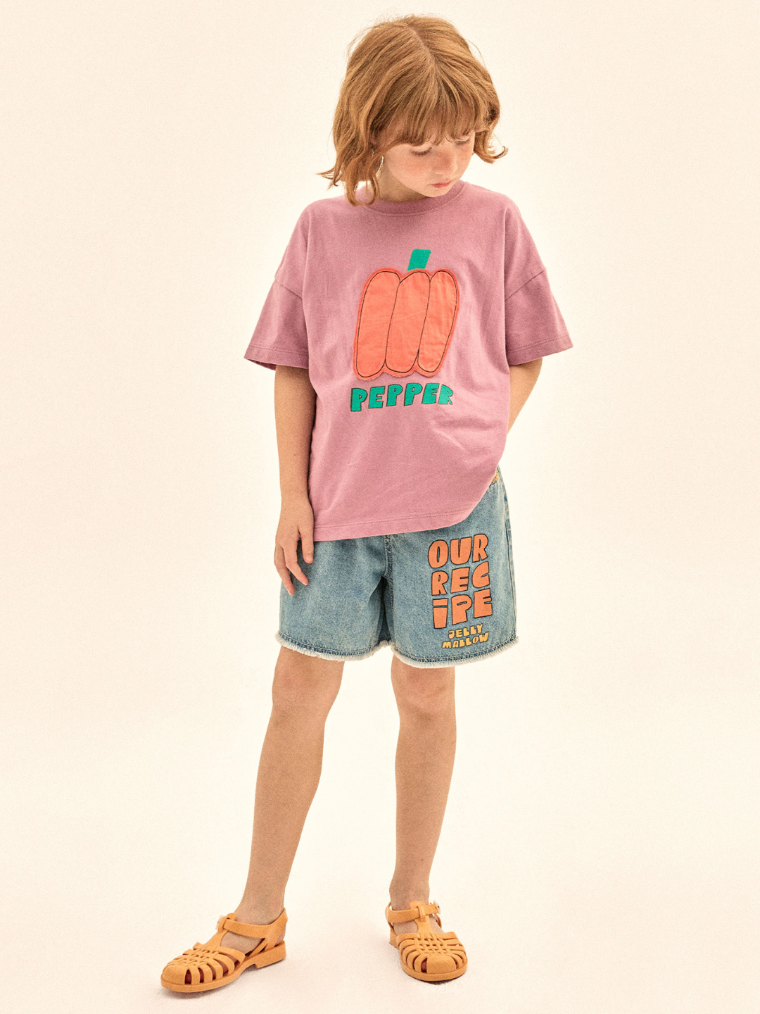 Child wearing a pink t-shirt with a red pepper design and denim shorts on a beige background
