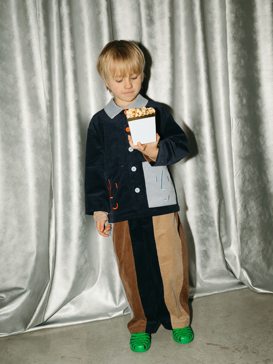 Second image of Navy Blue | The FACE JACKET is a navy blue kids corduroy jacket with a light blue collar, mismatched buttons, and two large front pockets embroidered with red and light blue smiling faces.