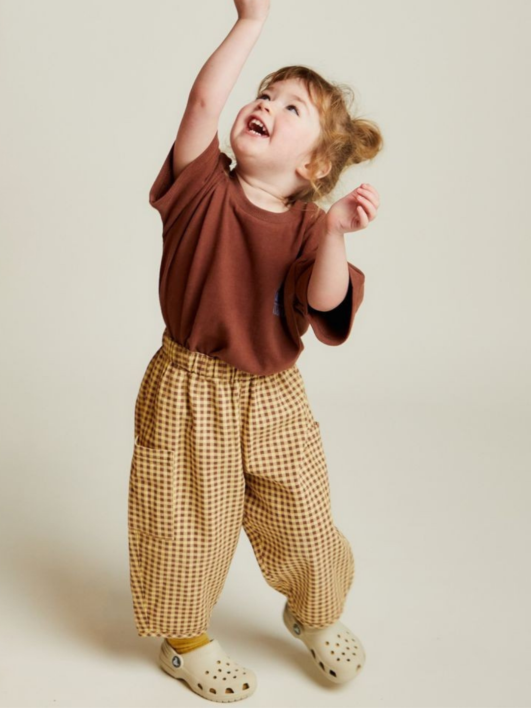 A young child smiles and looks up, reaching overhead while wearing EMERSON OCHRE GINGHAM TROUSER, a brown shirt, and beige clogs.