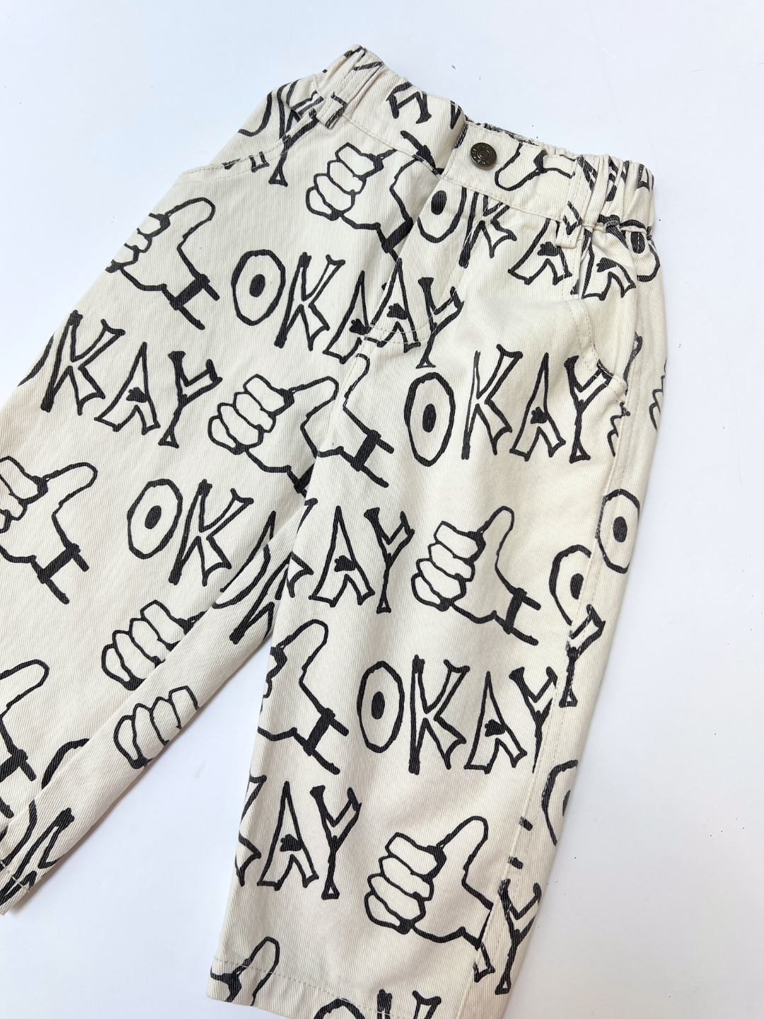 OKAY JEANS: Relaxed-fit beige denim pants featuring black "OKAY" text and thumbs-up hand graphics printed all over.