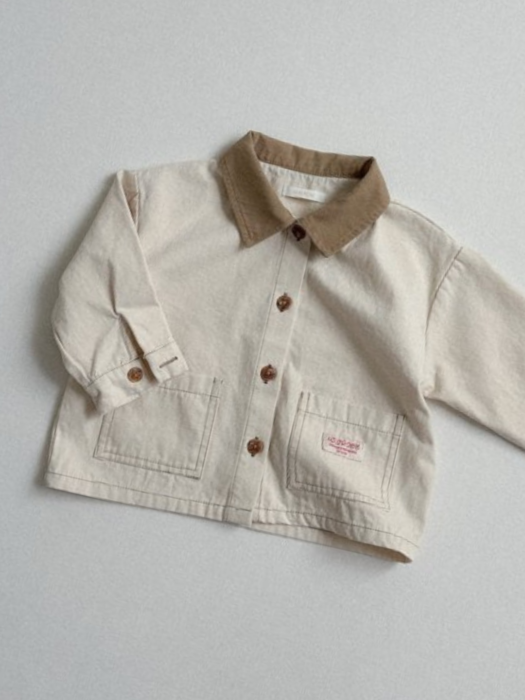 Ecru | The MINI WORKWEAR JACKET, a beige kids’ jacket with a brown collar, brown buttons, and two front pockets, is displayed on a light gray surface.