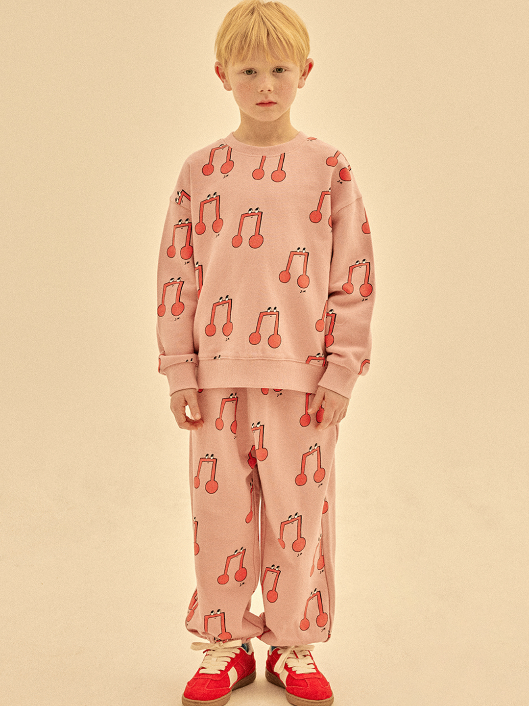 A young child wears the MELODY PATTERN SWEATSHIRT with matching pink pants featuring red cherries and red sneakers, standing against a beige background—this adorable look is simply irresistible.