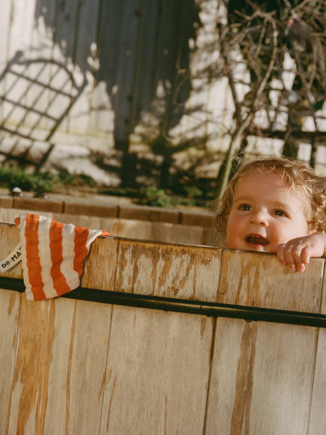 A young child with curly hair peeks over a wooden tub outdoors, where ORGANIC BABY WASH CLOTHS made of red and white striped muslin cotton are draped over the side.