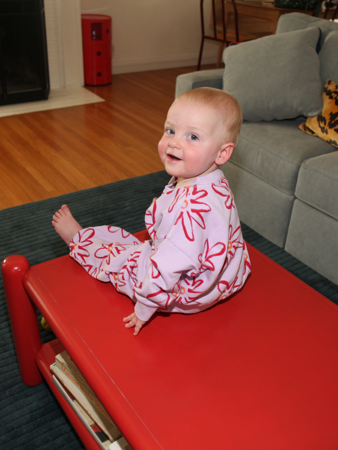 Lilac | A baby wearing the CHARLIE SWEAT SET with a painted flower print made from organic cotton sits on a red table in the living room, smiling and looking back over their shoulder.
