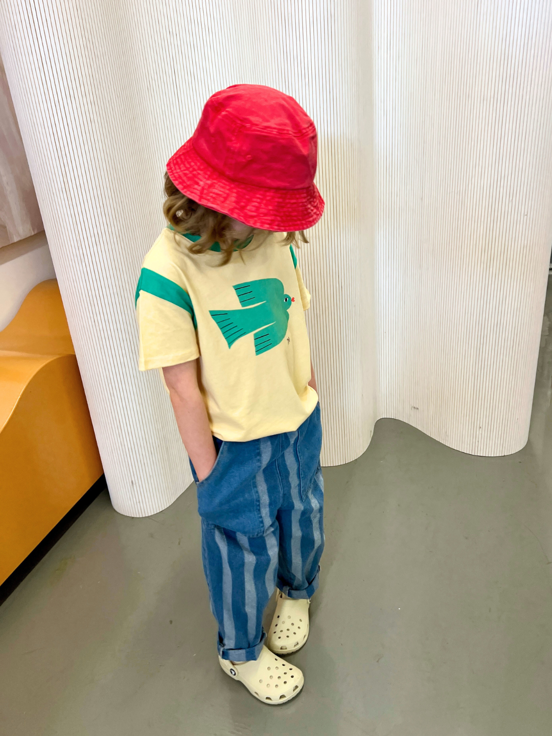 Red | A child wearing the PIGMENT BUCKET HAT in red, with a cream t-shirt with a green bird, and striped blue jeans, in an art studio.