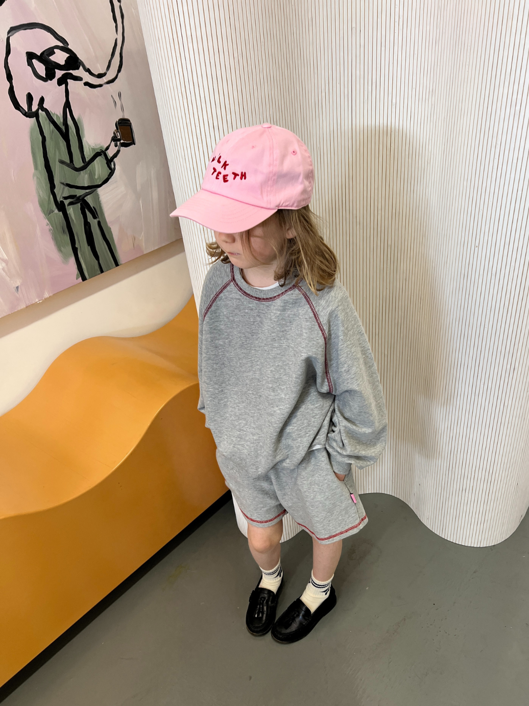 Pink | Child wearing the Milk Teeth baseball cap in bubblegum pink with red Milk Teeth lettering, with a grey sweatshirt and shorts and black loafers and white socks, inside an art studio.