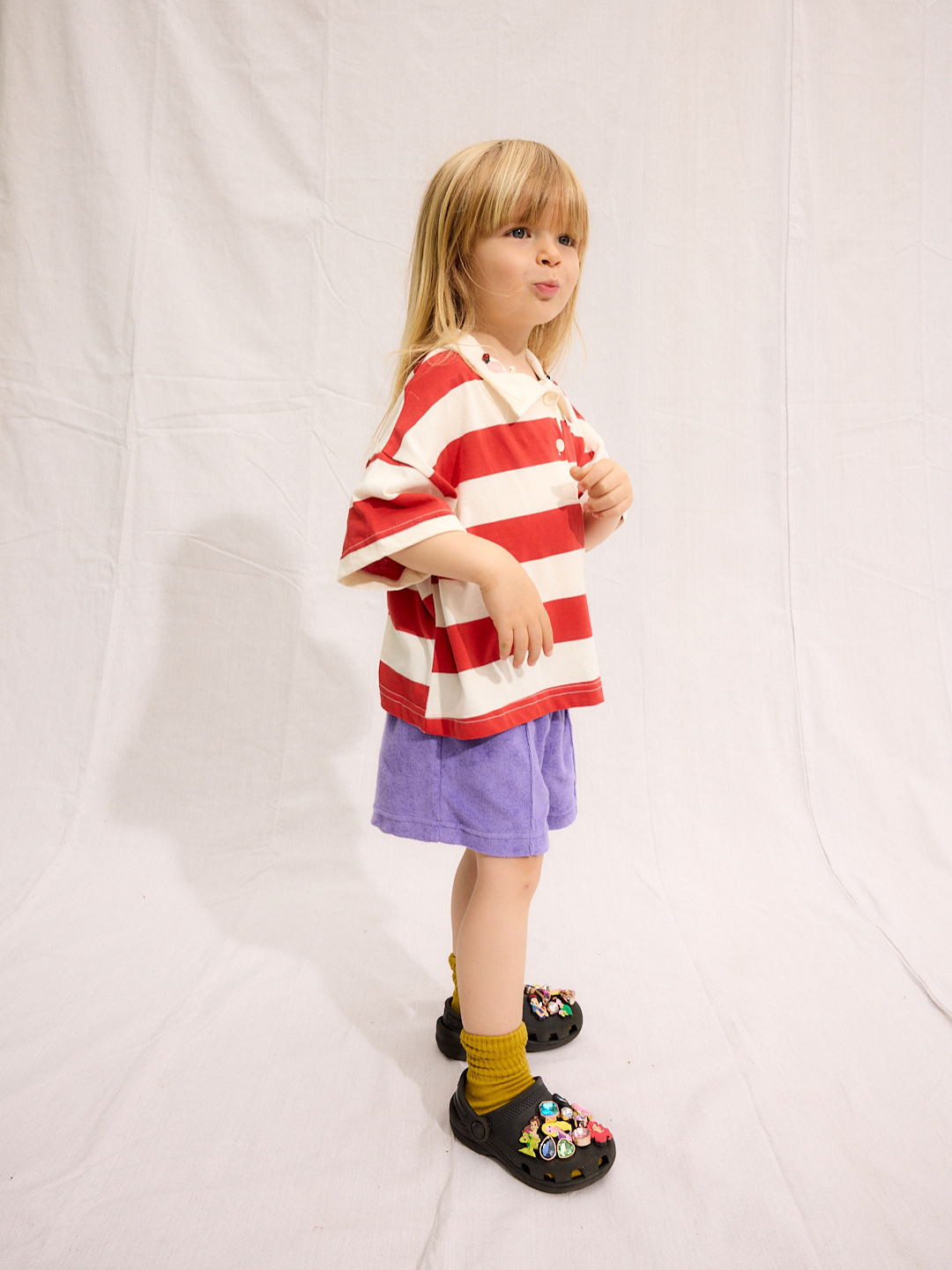 A young child stands against a white backdrop, wearing the RUGBY COLLAR TEE—a 100% cotton red and white striped shirt from a Korean kids brand—paired with purple shorts, yellow socks, and black clogs decorated with colorful charms.