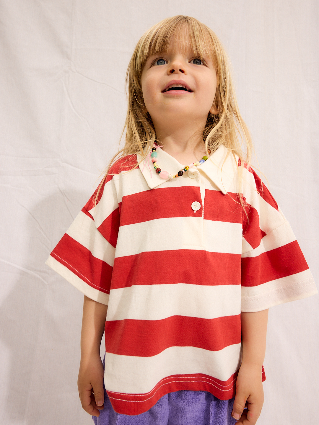 A young child with long blond hair wears the RUGBY COLLAR TEE, a 100% cotton, lightweight red and white striped shirt from a Korean kids brand, paired with a beaded necklace and purple shorts against a plain white background.