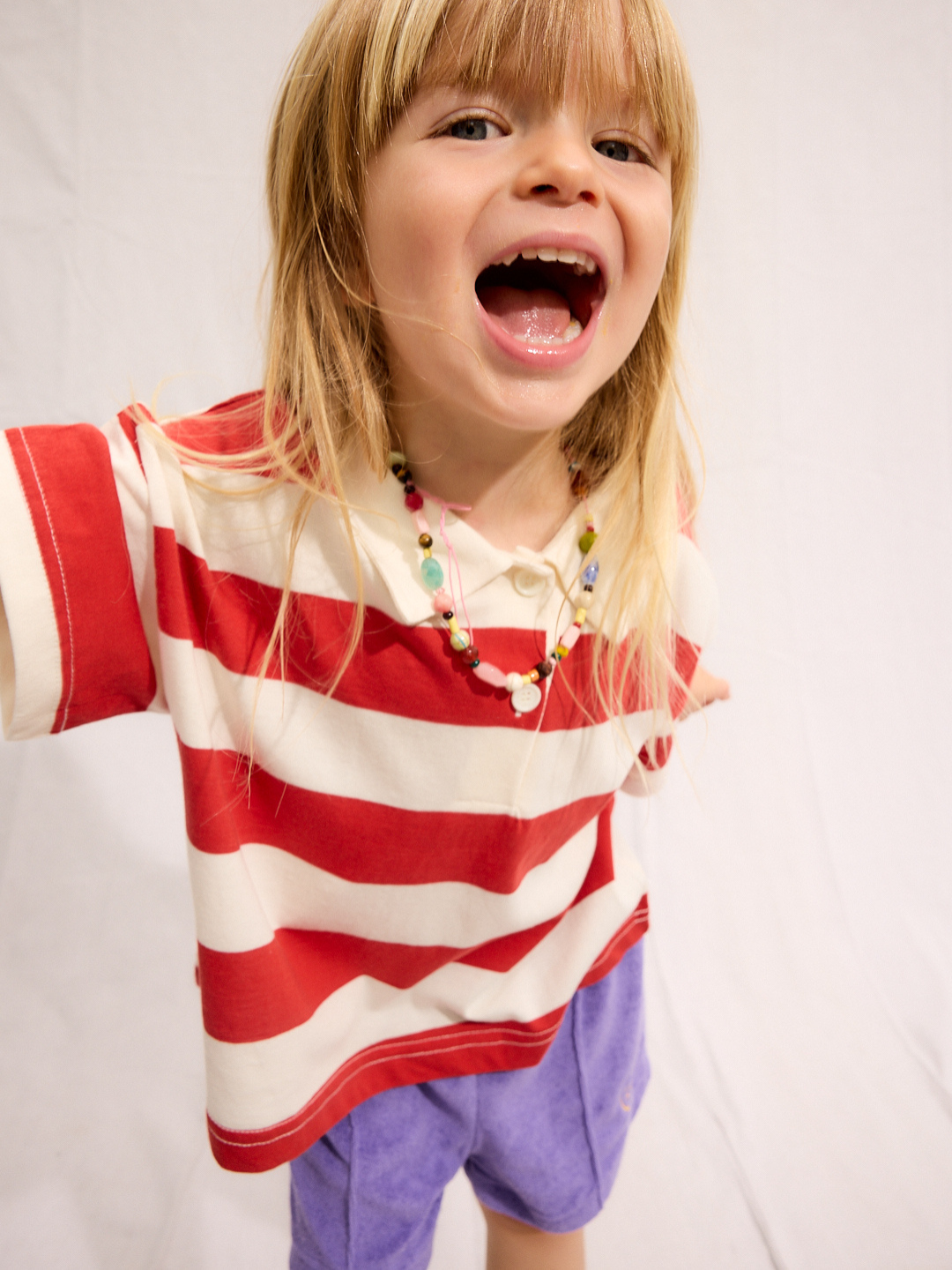 Young child with long blonde hair smiles with mouth open, wearing the RUGBY COLLAR TEE—an eye-catching red and white striped 100% cotton shirt from a Korean kids brand—along with purple shorts and a colorful beaded necklace against a plain background.
