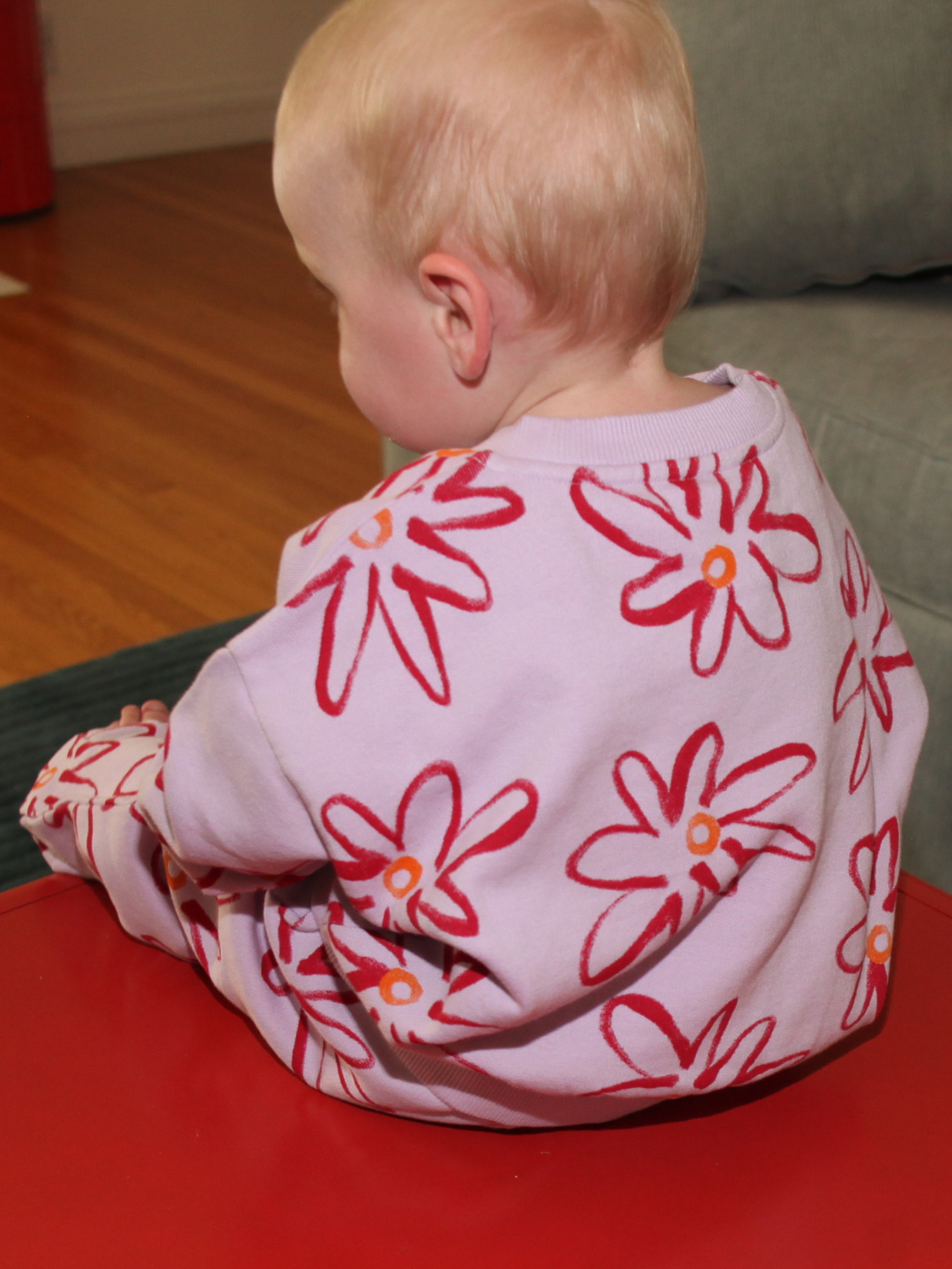 Lilac | A small child with short blonde hair sits on a red table, facing away, wearing the CHARLIE SWEAT SET—an organic cotton sweat set in light purple with a red and orange painted flower print.