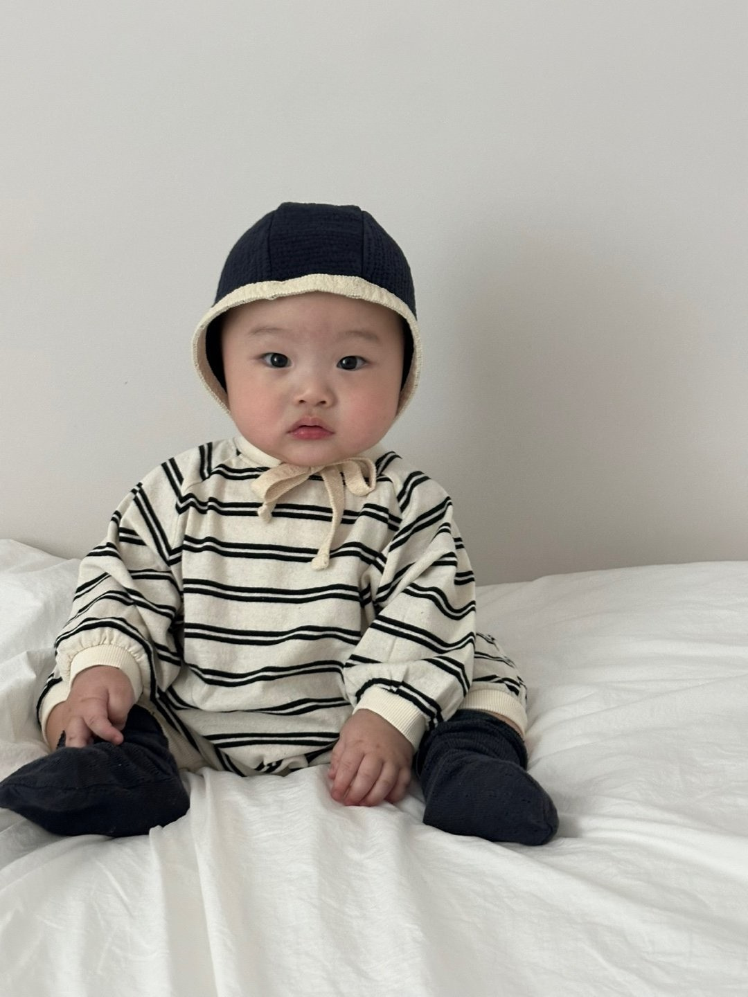 Off-White | A baby wearing the SLOUCHY STRIPE ROMPER from a Korean baby brand sits on a white bed, accessorized with a bonnet and looking at the camera.