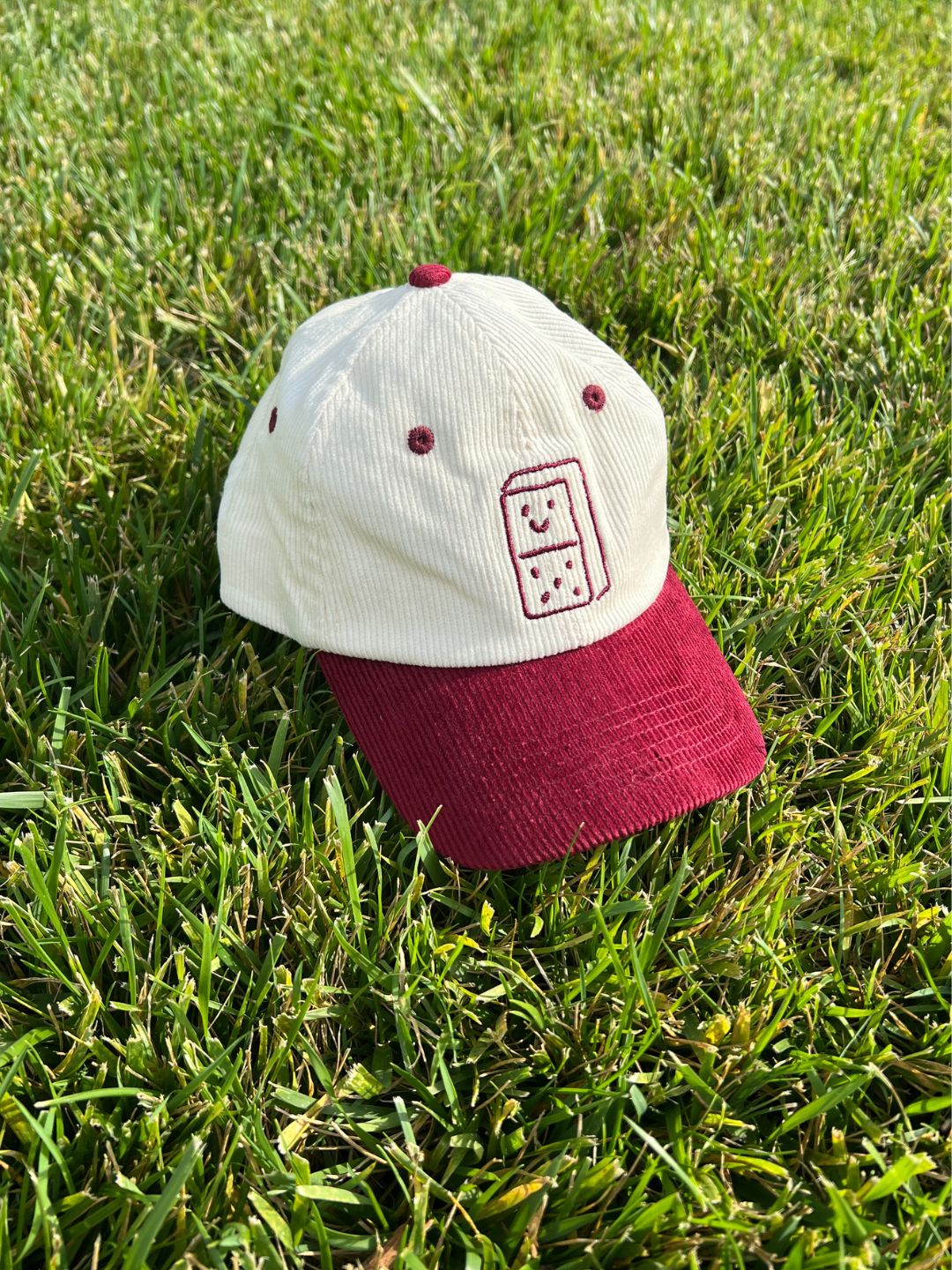 Red | A two-tone baseball cap resting on green grass. The crown is off-white corduroy with a small embroidered domino graphic on the front, and the curved brim is burgundy corduroy.