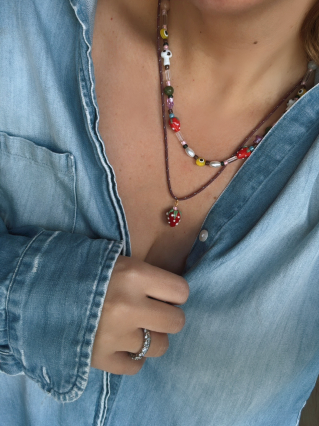 A person wears a light blue denim shirt slightly open at the chest, showcasing the GAIA NECKLACE—genderless jewelry with handmade strawberry and mushroom beads—and a silver ring on their finger.