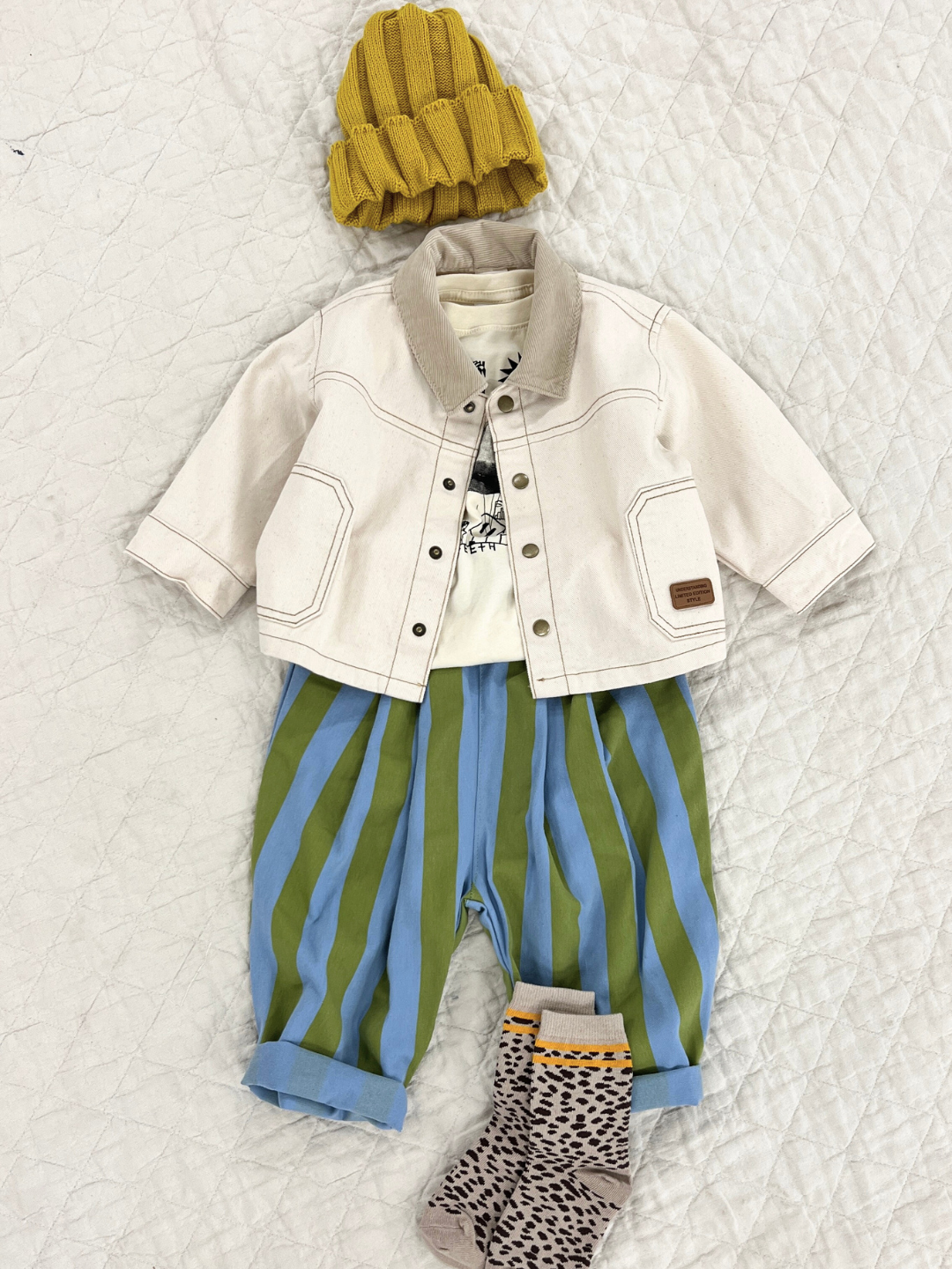 Natural | Front view of kids ecru denim jacket with beige corduroy collar. Jacket is shown open over a white t-shirt, and paired with a blue and white striped pant, a yellow beanie, and cheetah print socks.