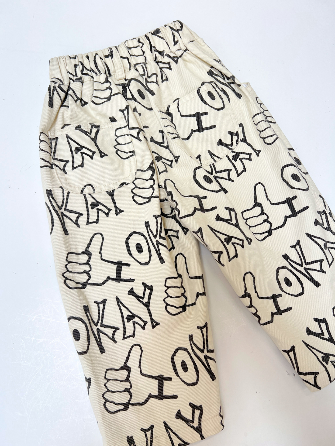Ecru | OKAY JEANS: Beige relaxed-fit jeans with black "OKAY" text and a thumbs-up hand graphic, shown flat on a white background.