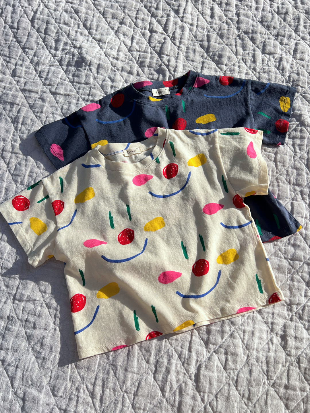 Cream | Two kids' Facepaint Tees in Navy and cream laid flat on a quilt in the sun