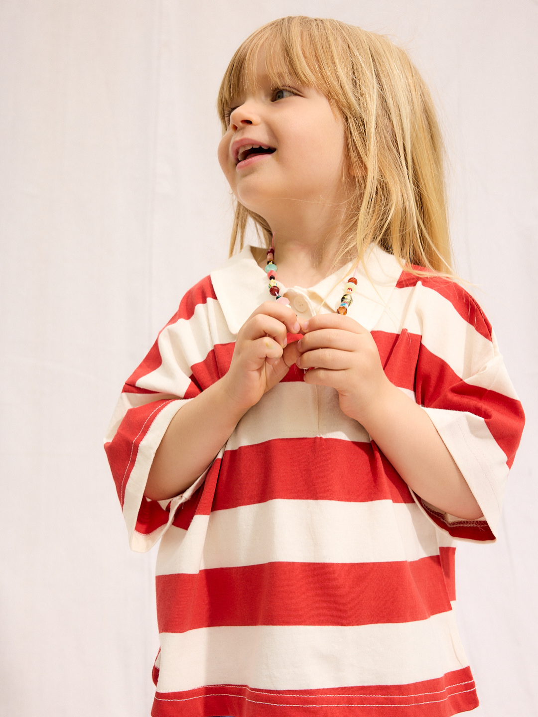 A young child with long blonde hair smiles and looks up while wearing the RUGBY COLLAR TEE, a lightweight 100% cotton red and white striped shirt from a trusted Korean kids brand, paired with a beaded necklace against a plain light background.