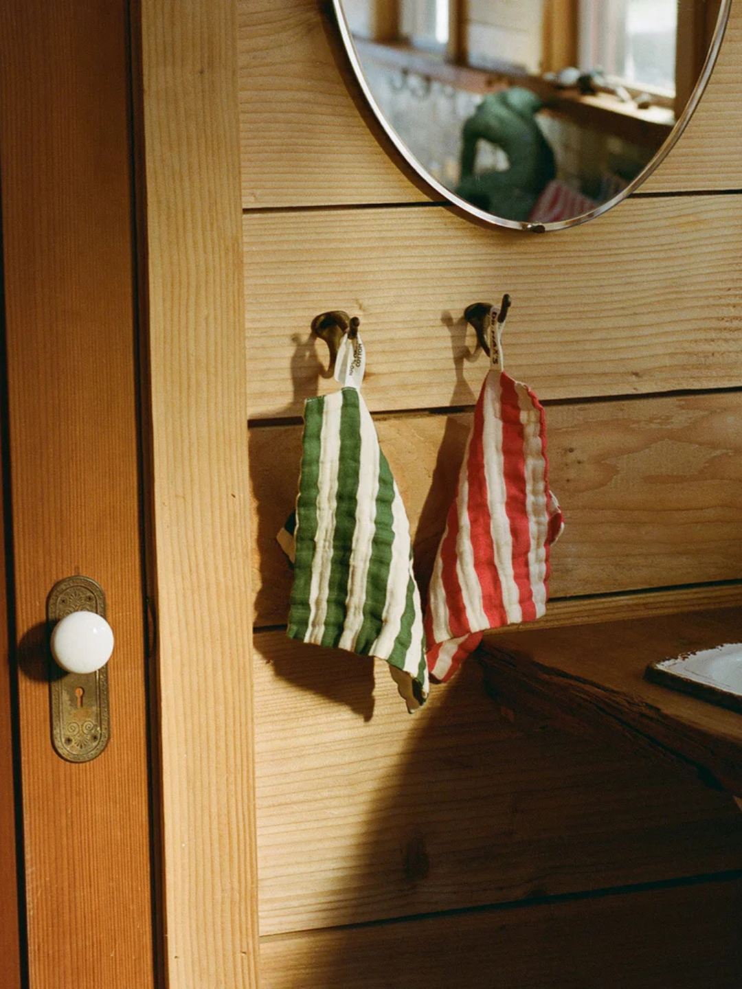 Two ORGANIC BABY WASH CLOTHS—one green, one red—hang on hooks below a round mirror on a wooden wall beside a door with a white knob.