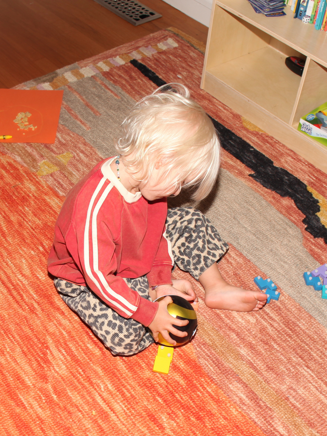 Red | A young, blonde-haired child sits cross-legged on a patterned rug, wearing the oversized HALFTIME LONGSLEEVE and leopard print pants, playing with a toy ball and blue plastic animals.