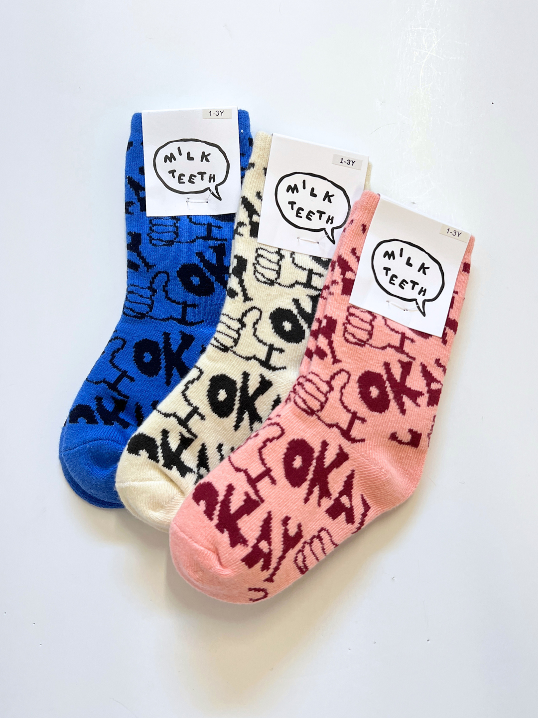 Pink | The OKAY SOCK set includes three pairs of children's socks in blue, cream, and pink, each featuring hand-drawn "OK!" and thumbs-up patterns on stretchy cotton for comfort—labeled as "Milk Teeth.