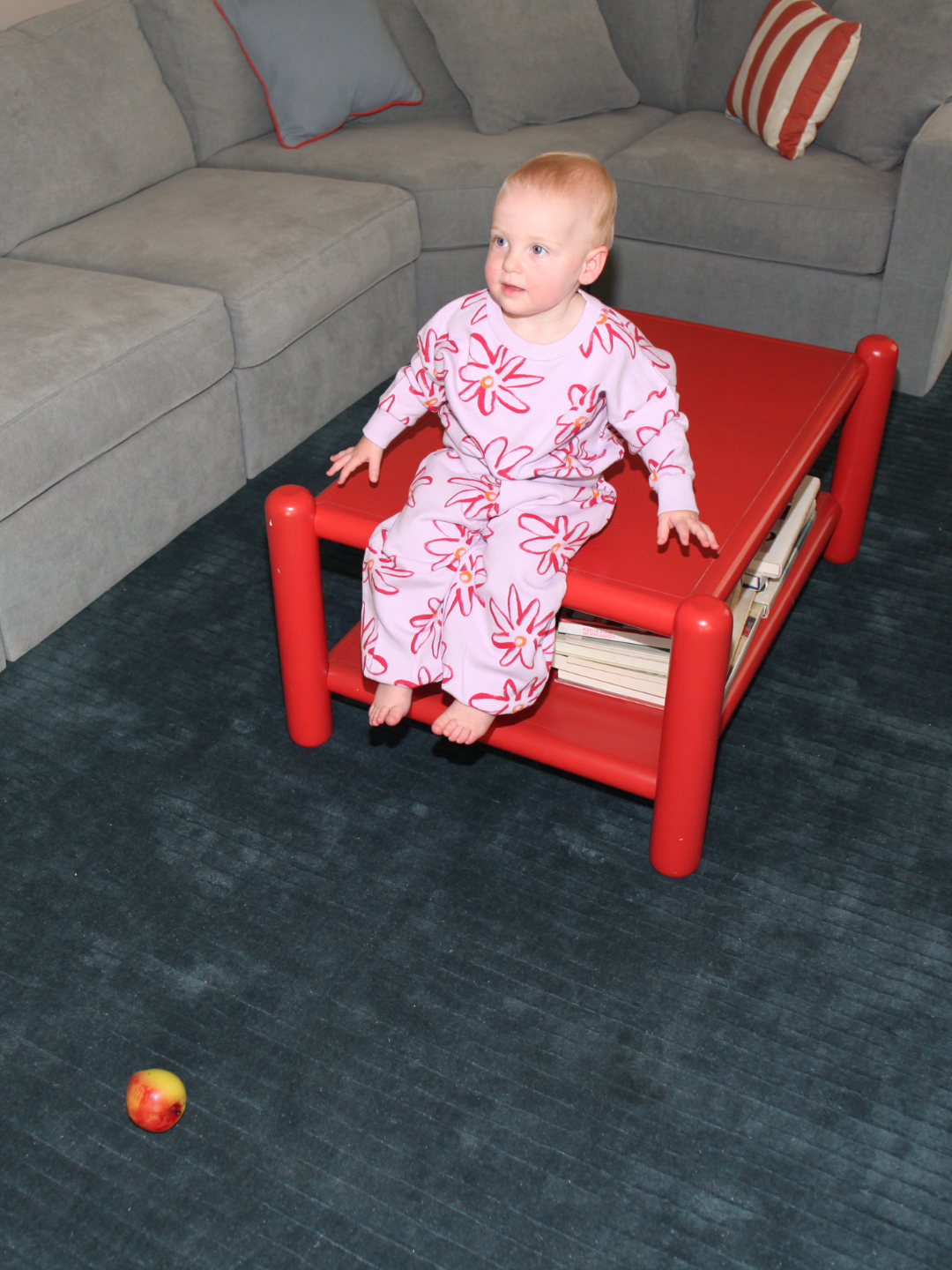 Lilac | A baby wearing the CHARLIE SWEAT SET in pink floral French Terry sits on a red table in a living room with a gray couch, gazing at an apple on the dark carpet.
