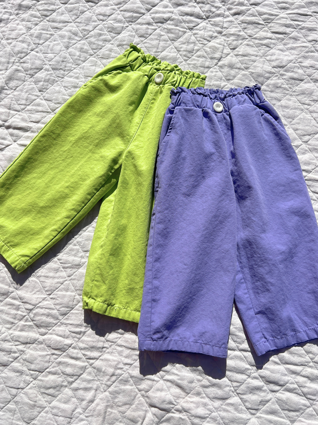 Purple | Two pairs of JELLY PANTS, children's shorts crafted from soft woven cotton in green and purple, lay on a quilted white surface. Each pair features an elastic waistband and button, embodying the relaxed pull-on style typical of a top Korean kids brand.