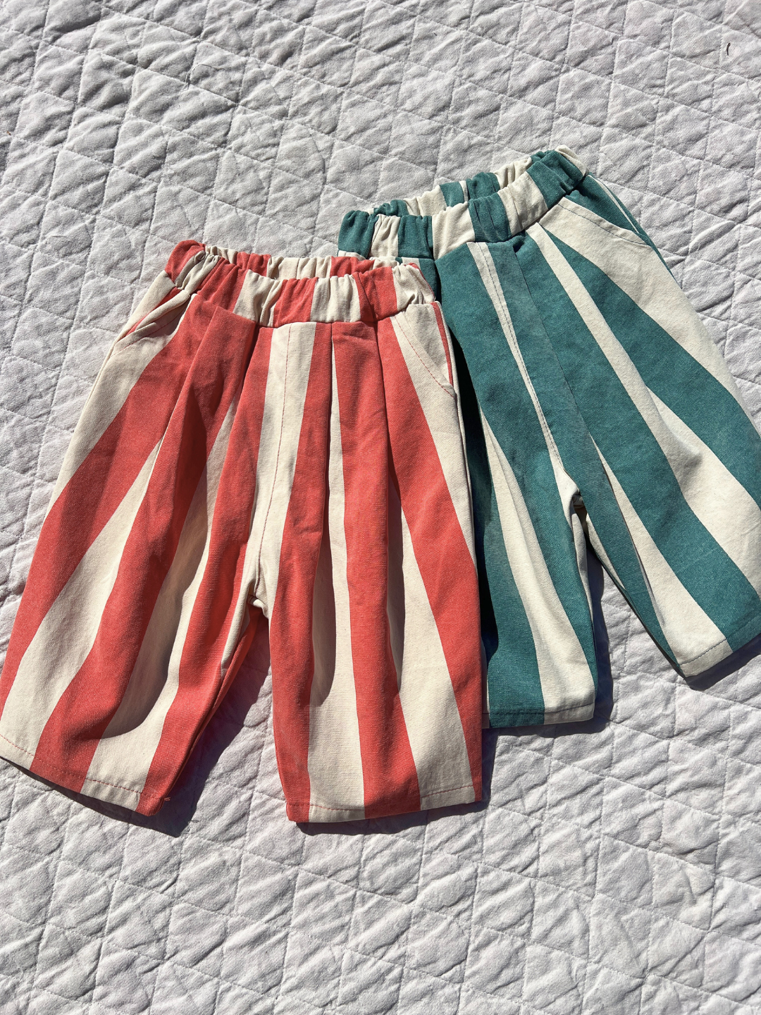 Faded Red | Two pairs of BIG STRIPE PANTS in red/white and green/white stripes rest on a quilted surface. Crafted from lightweight cotton canvas, they're ideal for warm days.