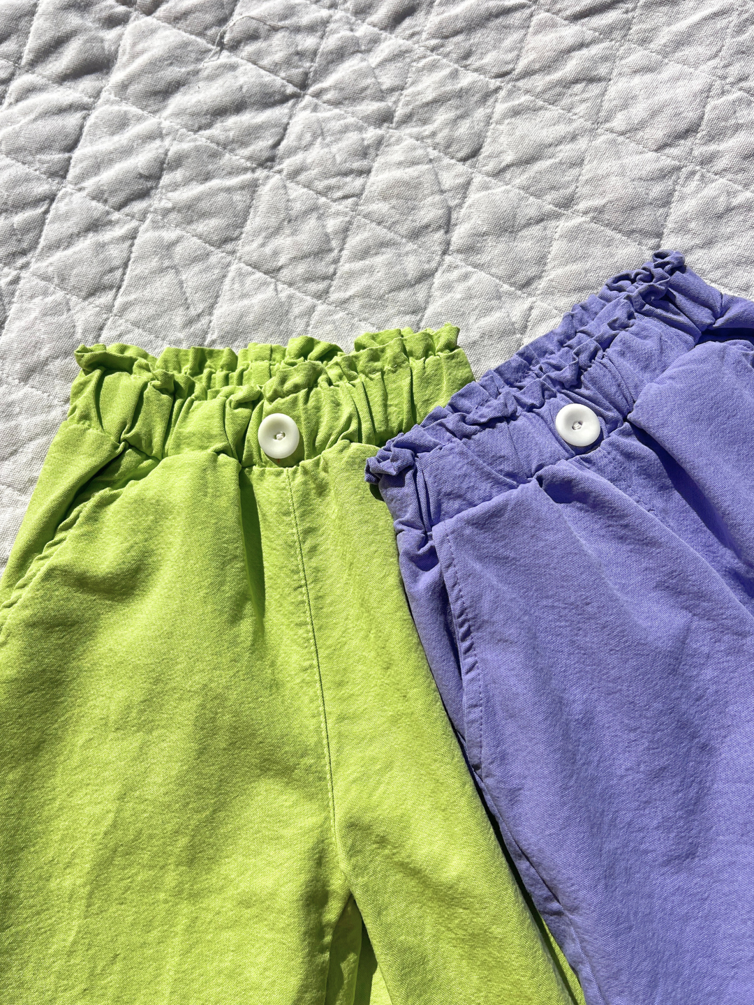 Purple | Two pairs of JELLY PANTS, one green and one purple, rest on a quilted surface. Both have ruffled elastic waistbands with white buttons, crafted from woven cotton by a Korean kids brand.
