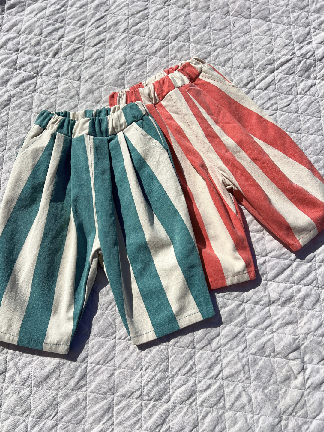 Teal | Two pairs of BIG STRIPE PANTS for children, one blue and white, the other red and white, made from lightweight cotton canvas and showcased on a quilted surface.