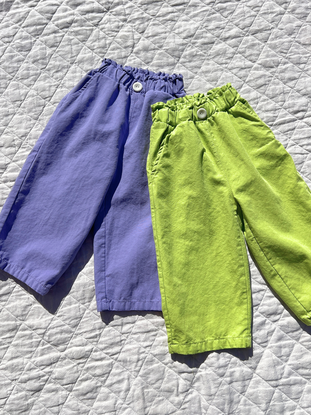 Green | Two pairs of JELLY PANTS, crafted by a Korean kids brand, are laid on a quilted surface. One pair is purple and the other light green, both with elastic waistbands and buttons for easy pull-on wear. Made from soft woven cotton for comfort.