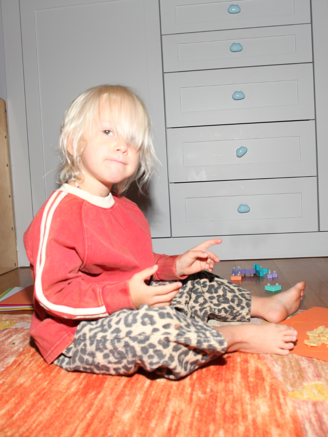 A young child with blonde hair sits on an orange rug, wearing the HALFTIME LONGSLEEVE in an oversized fit with leopard-print pants, surrounded by small toys and puzzle pieces.