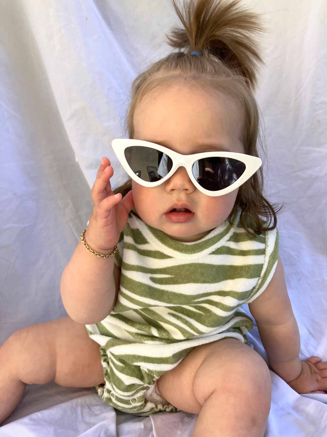 A child wearing the white Stingray kids Sunglasses. She is seated on a beige cloth backdrop and wears a beige terrycloth romper with sage green tiger stripes, with her hair in a top ponytail.